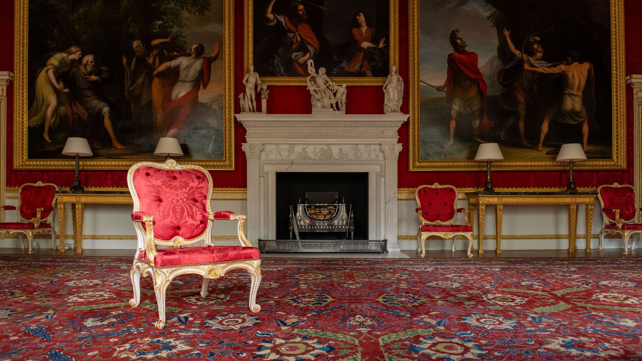 An empty gilt armchair upholstered in red silk damask, set before a marble fireplace beneath old master paintings