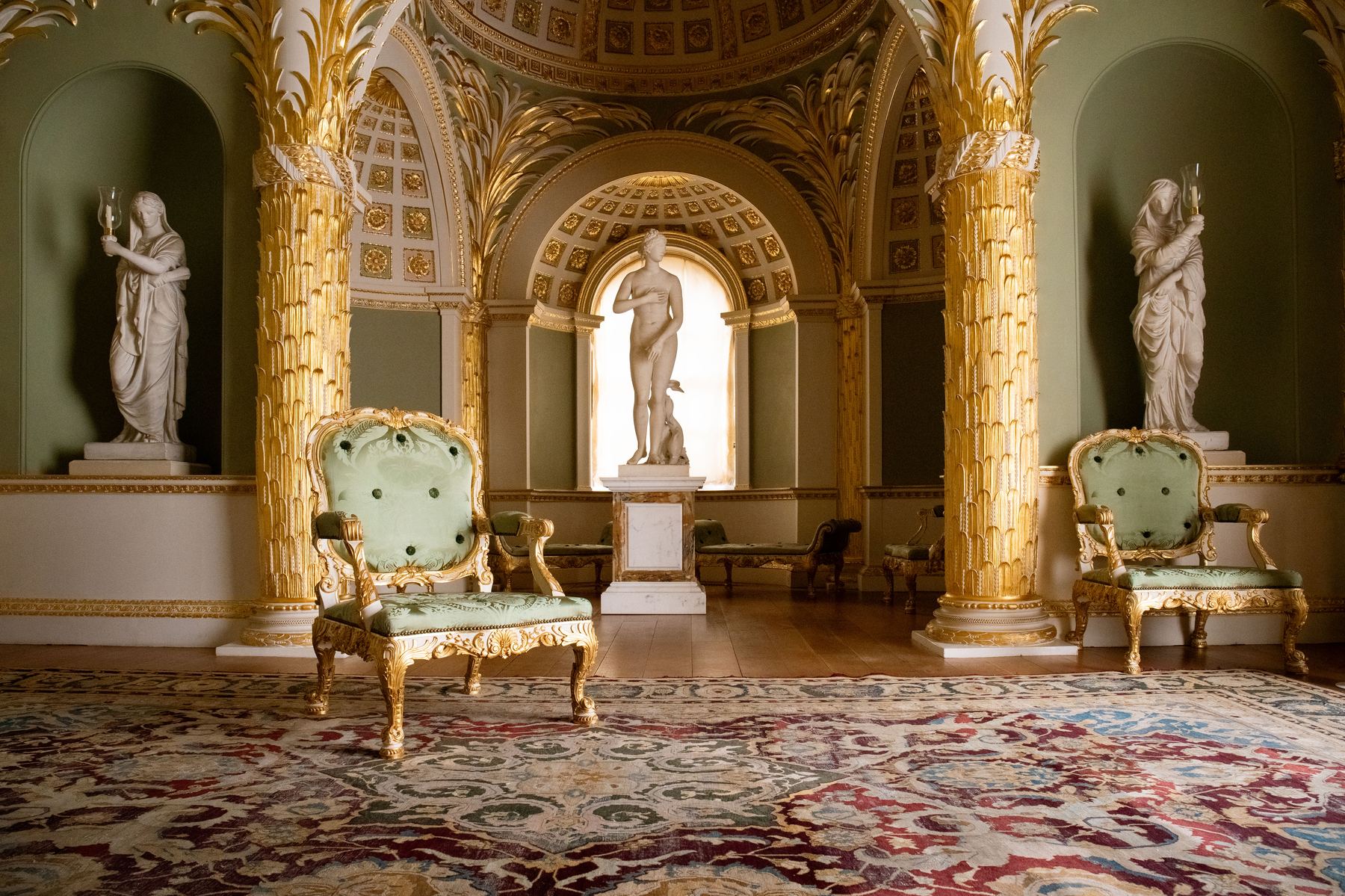 An empty green silk armchair between gilded palm columns, beneath a coffered dome