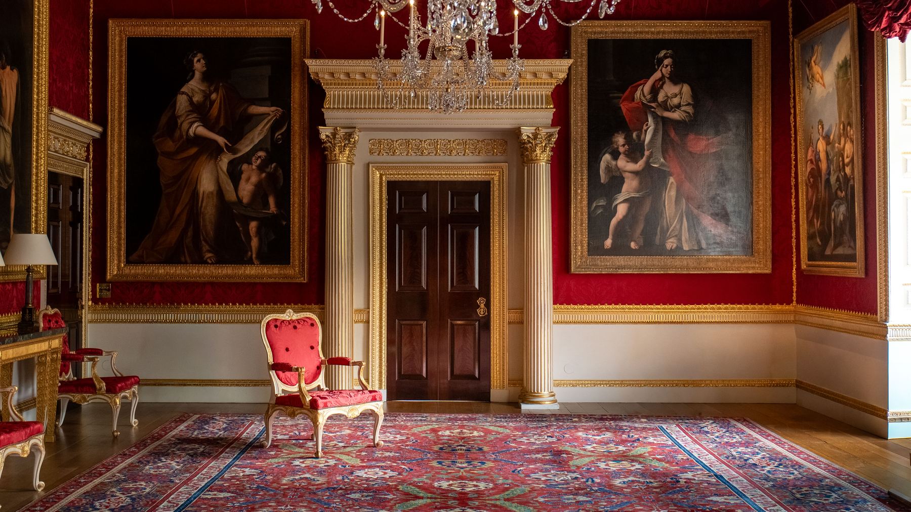 Red damask drawing room with gilt chairs, crystal chandelier and a pedimented doorway